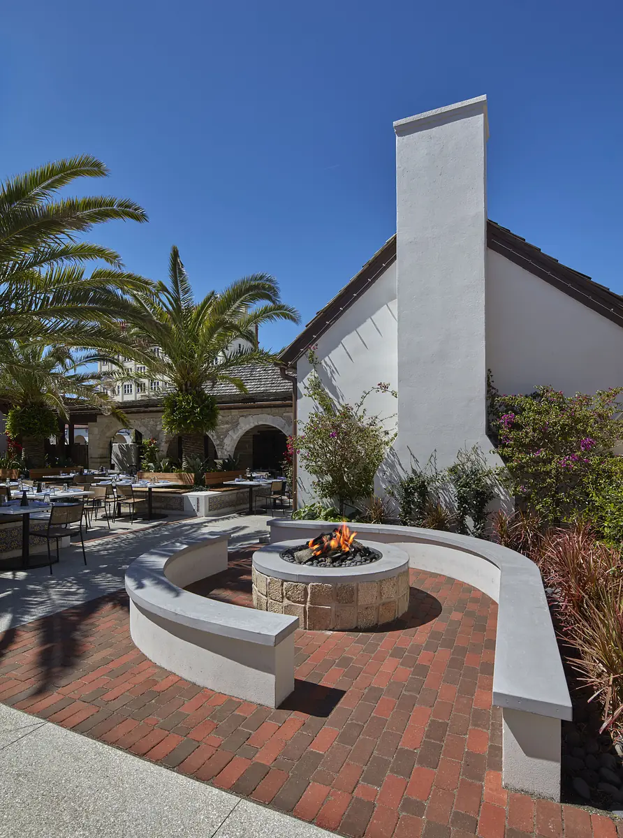 Outdoor patio with circular fire pit, concrete seating, brick flooring, and surrounding greenery.