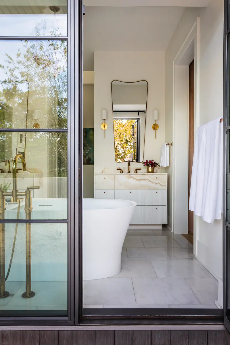 Bathroom with freestanding white tub, stone vanity, gold faucets, and wall sconces.