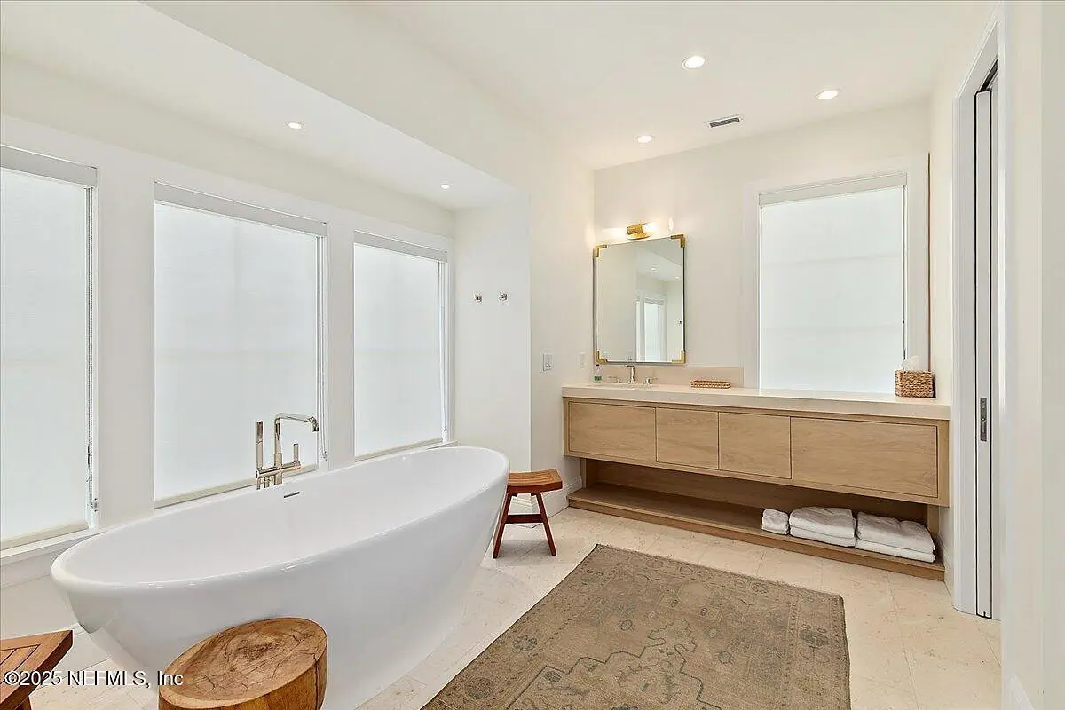 Bathroom with freestanding tub, wooden vanity, mirror, windows, and decorative area rug.