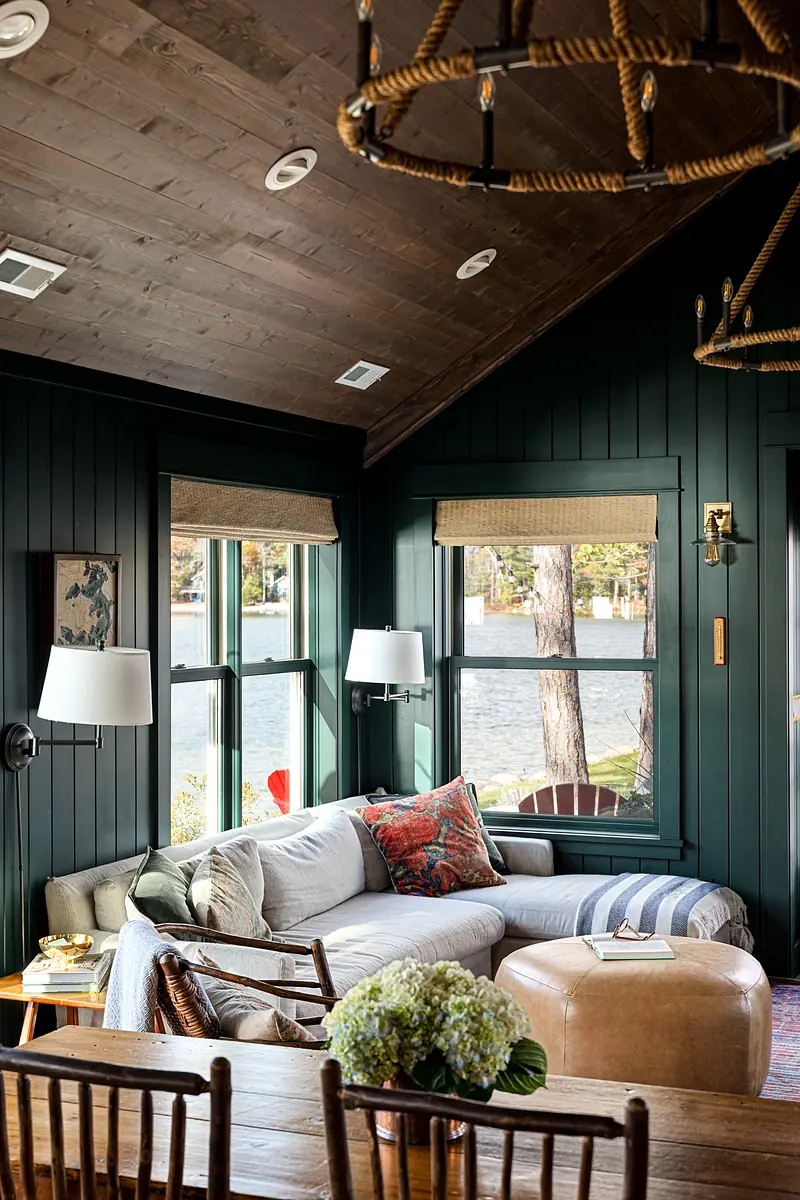 Living room with gray sectional, leather ottoman, wooden table, large windows, and dark green paneled walls.
