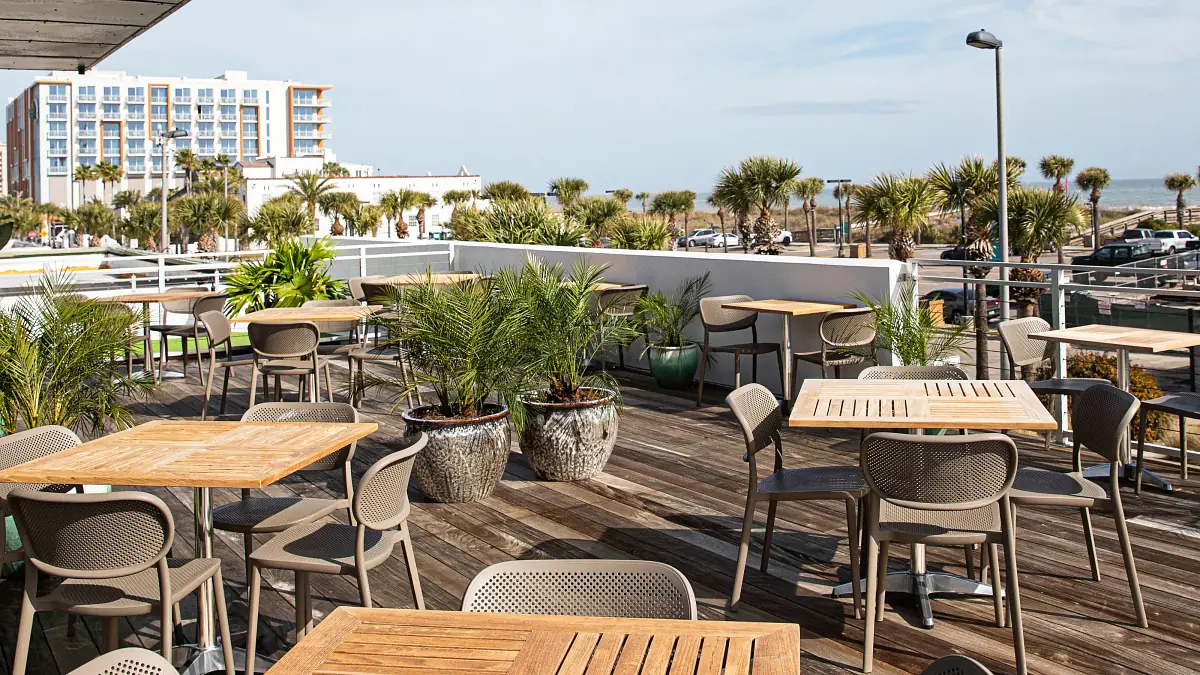 Outdoor patio with wooden tables, gray chairs, large potted plants, and wooden deck overlooking a skyline.