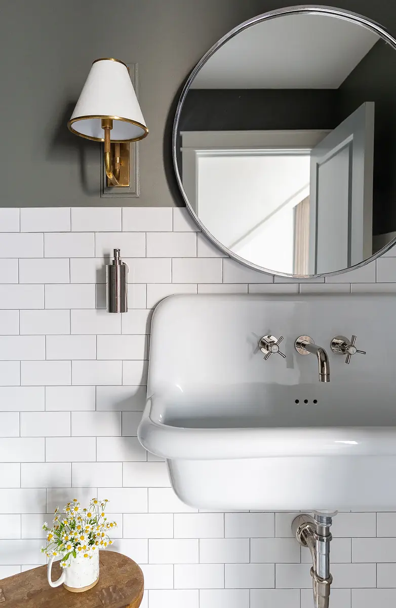 Bathroom with wall-mounted sink, round mirror, subway tiles, and wooden table with flowers