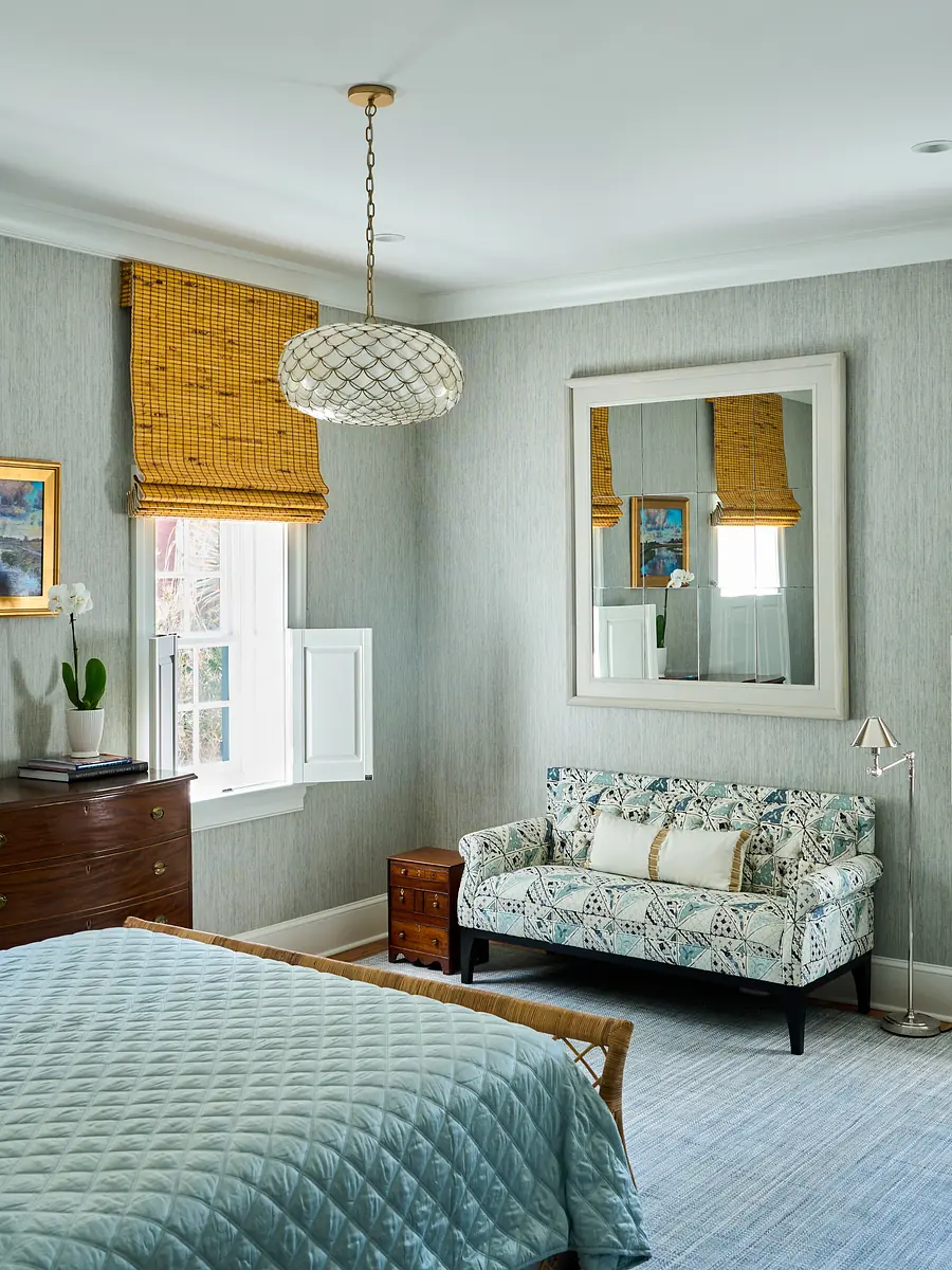 Bedroom with blue quilt, patterned settee, mirror, window with blinds, and wooden dresser.