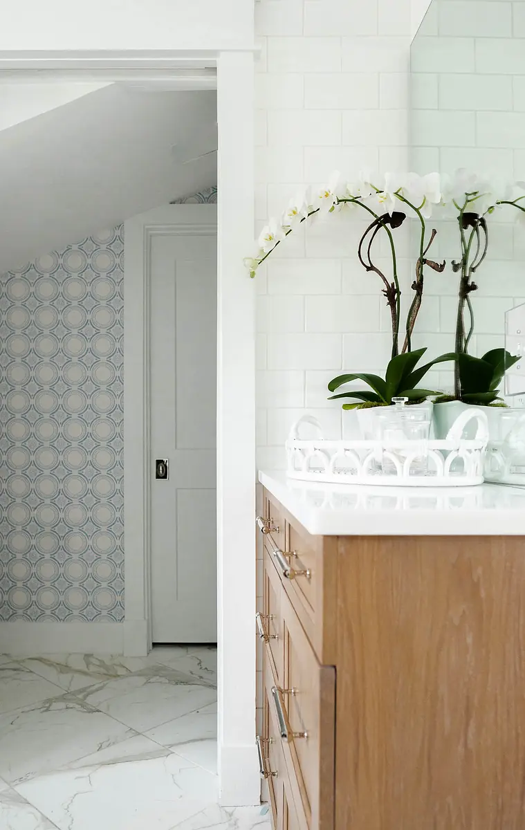 Bathroom with wooden vanity, white countertop, large mirror, patterned wallpaper, and potted orchid