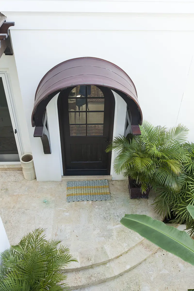 Exterior entryway with black door, brown arched overhang, potted plants, and welcome mat on stone floor.
