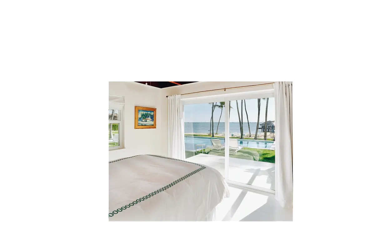 Bedroom with white bed, green trim, artwork, sliding doors, and view of palm trees and water.