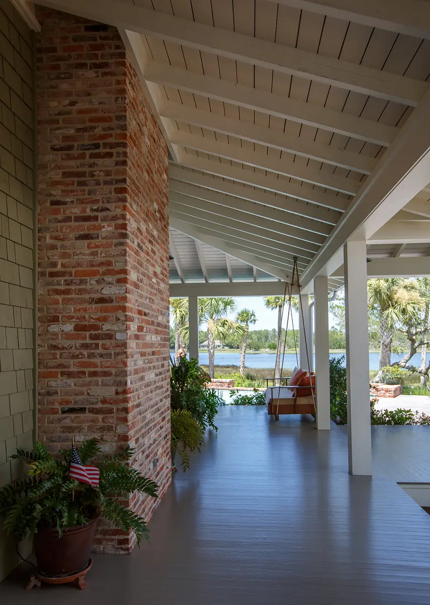 Patio with brick column, wooden swing, ferns, and views of water, featuring slanted ceiling with beams
