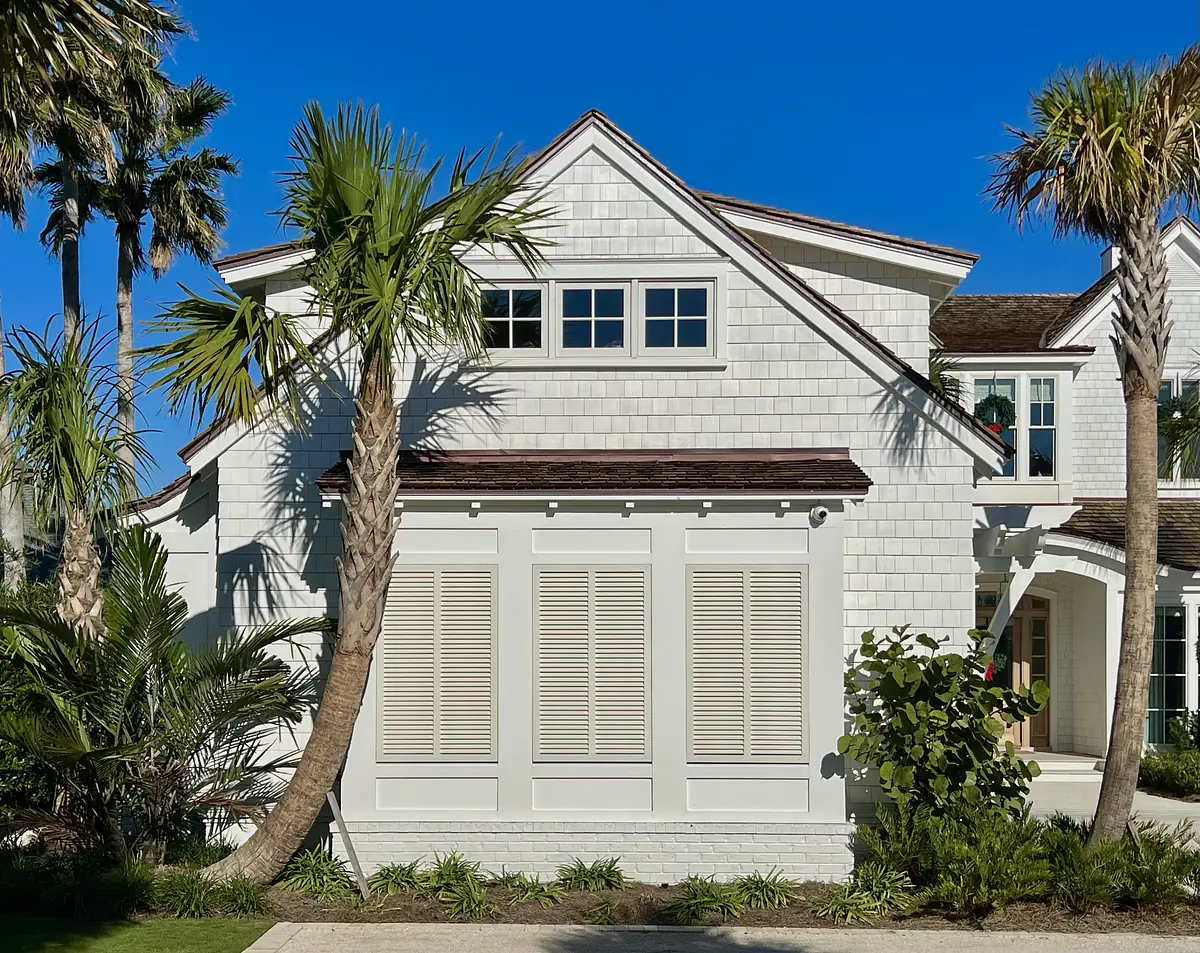 Exterior front of a house with white shingle siding, palm trees, windows, and landscaping.