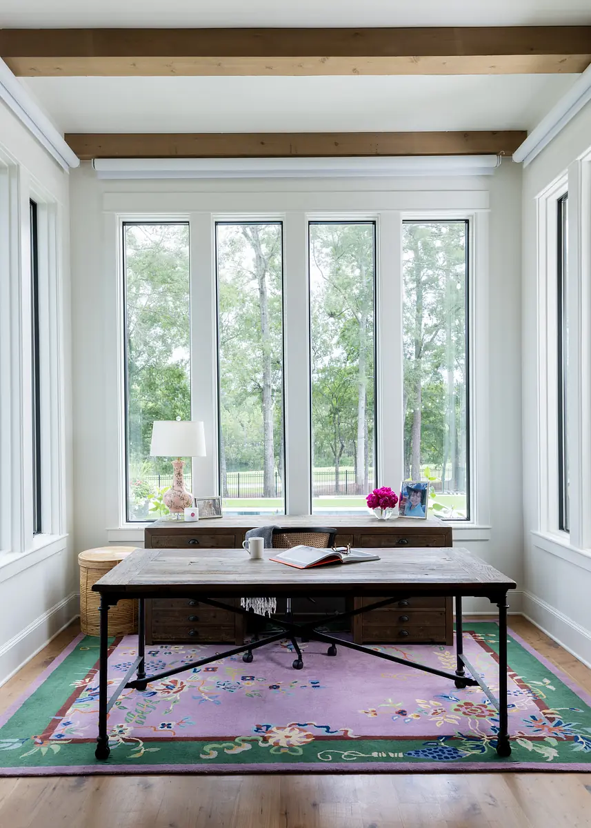 Office with wooden desk, white lamp, pink rug, storage unit, and large windows with outdoor views.