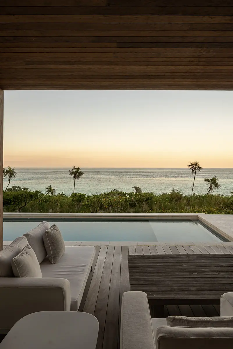 Patio with white sectional sofa, wood coffee table, swimming pool, and ocean view with palm trees.