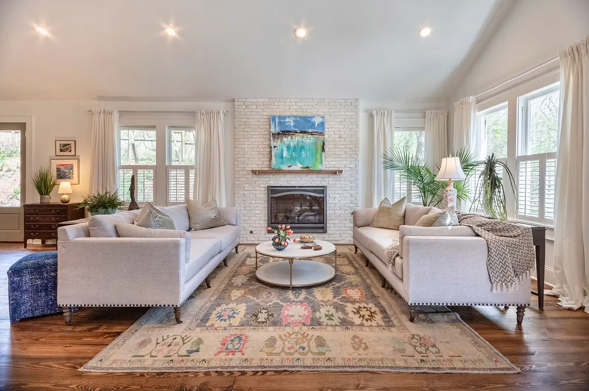 Living room with two gray sofas, round coffee table, area rug, white brick fireplace, and large windows.