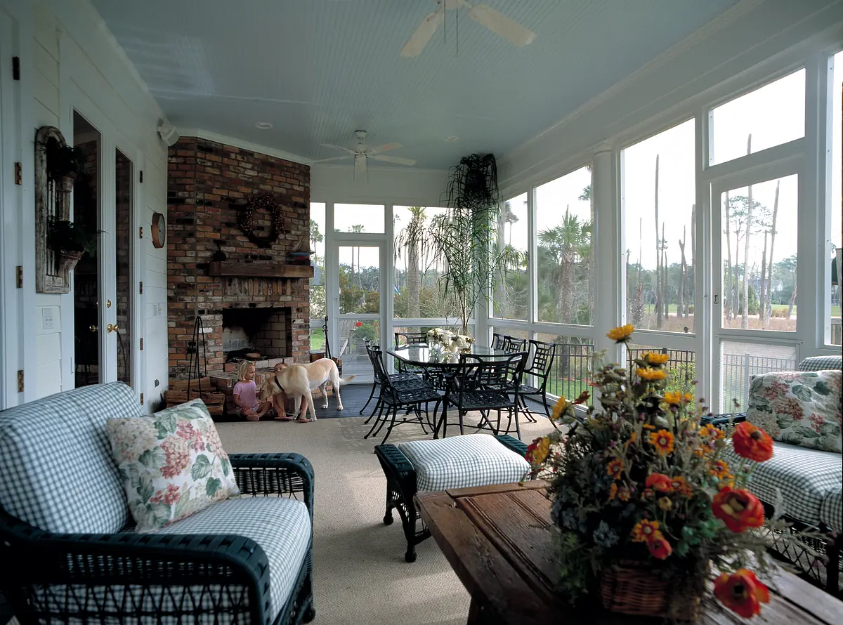 Sunroom with wicker sofas, wooden coffee table, glass dining table, brick fireplace, and large windows.