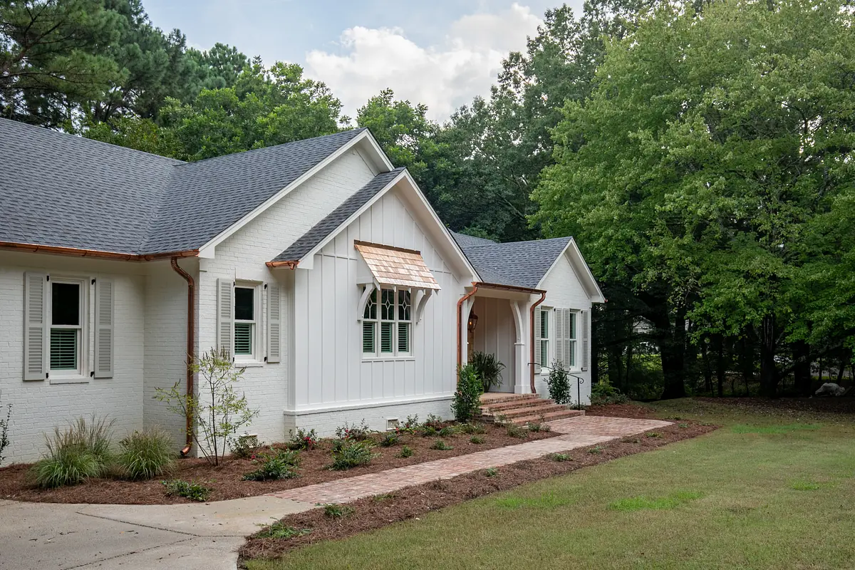 Front exterior of a house with white brick, wooden accents, landscaped shrubs, and a brick pathway.