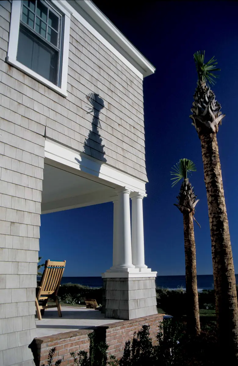 Exterior view of a house with wooden shingles, columns, rocking chair, palm trees, and ocean background.
