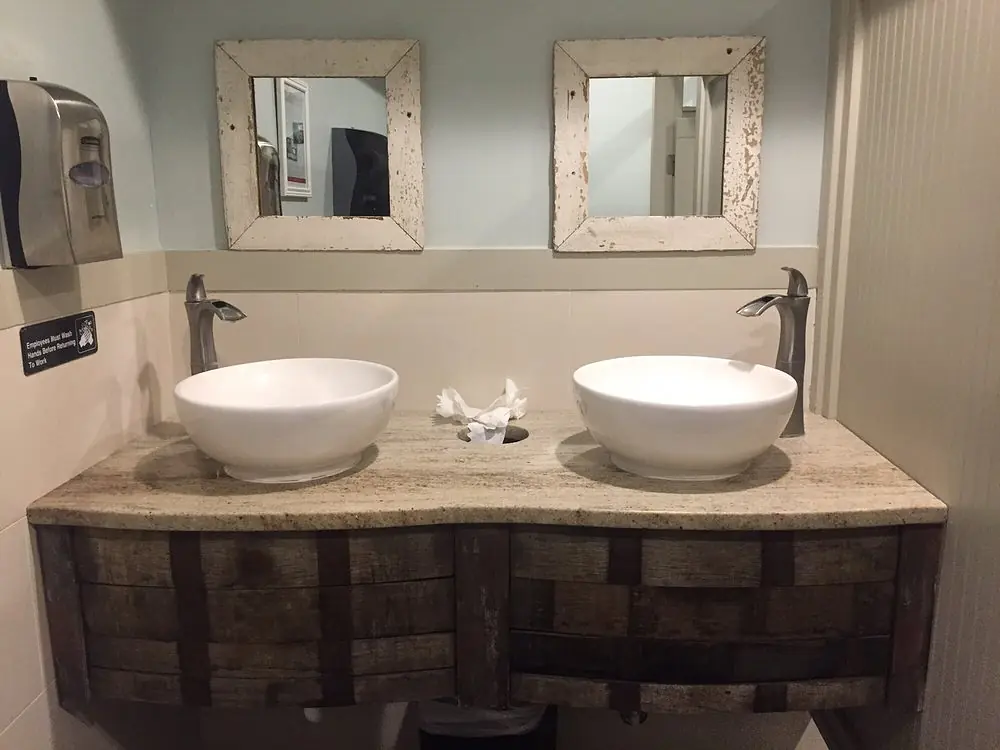 Bathroom with floating wooden vanity, white vessel sinks, rustic mirrors and light-colored tiled walls.