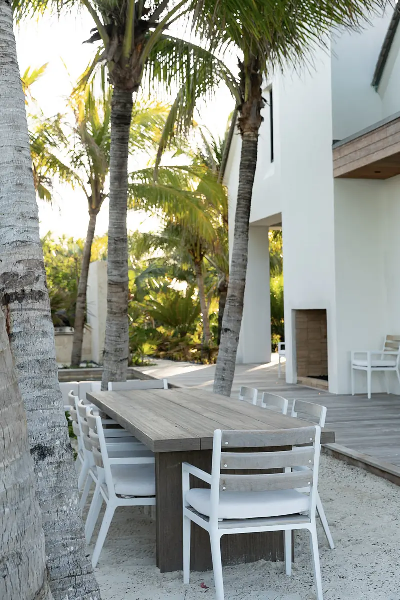 Outdoor patio with wooden dining table, white chairs, palm trees, and sand ground.