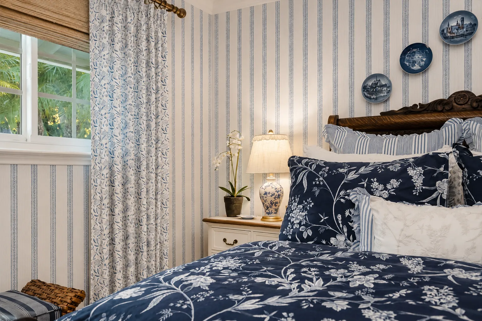 Bedroom with floral-patterned quilt, decorative pillows, nightstand, lamp, and striped walls.