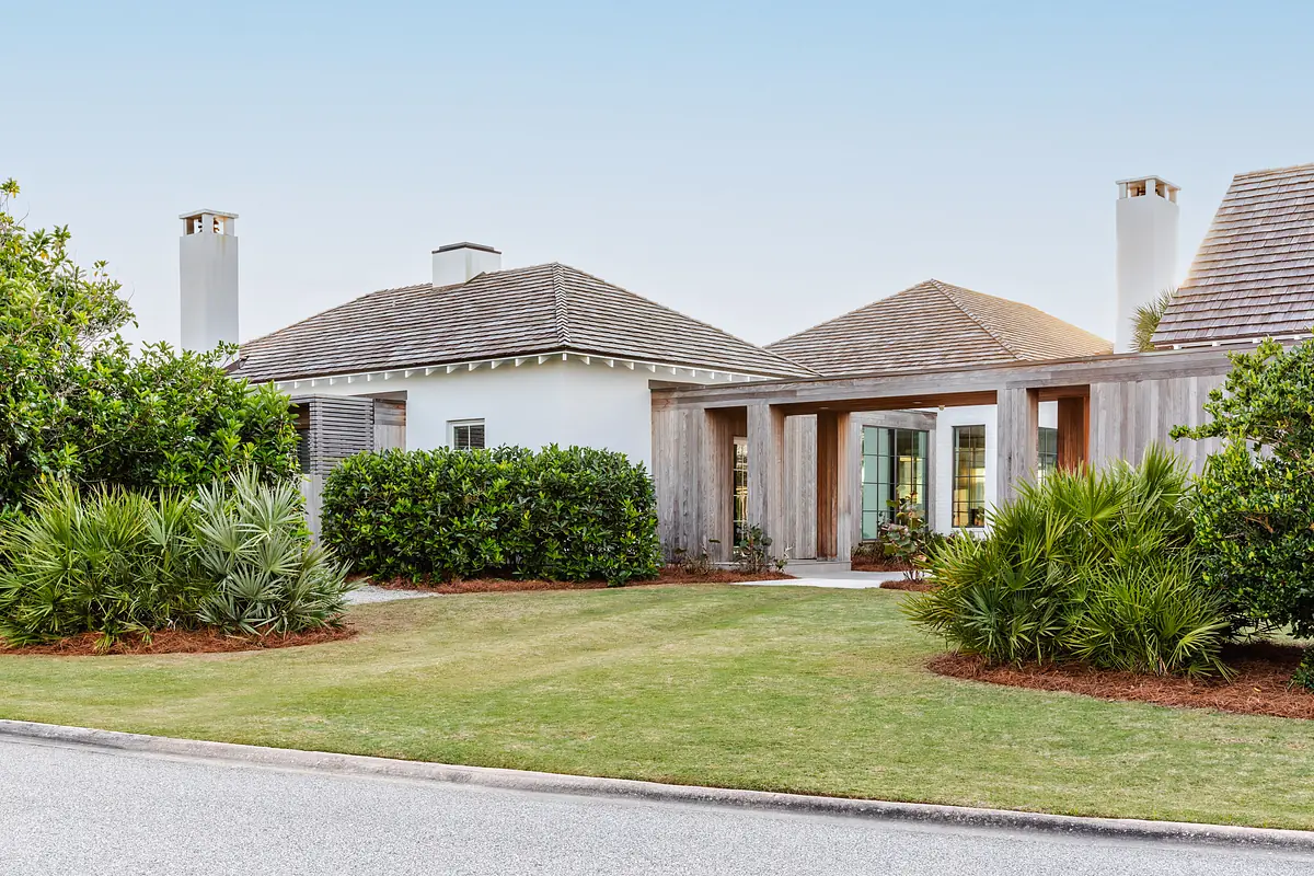 Front exterior of a house with sloped roof, chimneys, porch, shrubs, and palm plants on a grassy lawn.