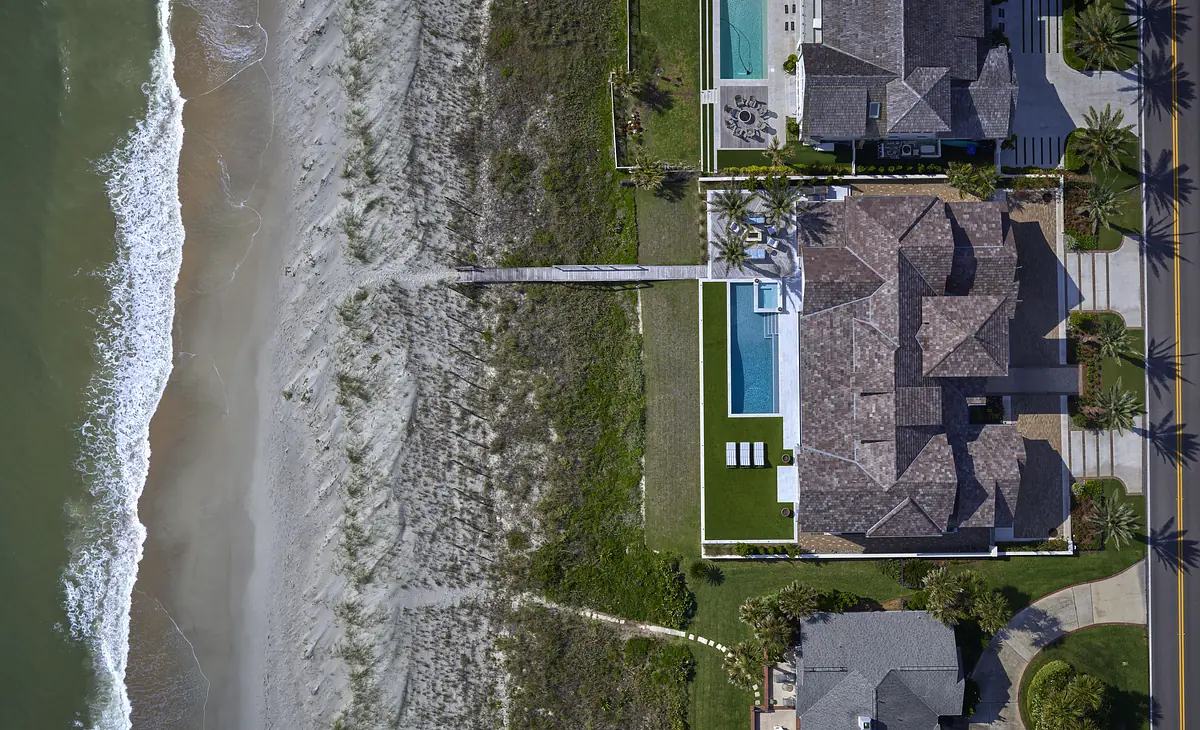 Exterior view of beachfront property with sandy shoreline, grass areas, swimming pool, and palm trees.