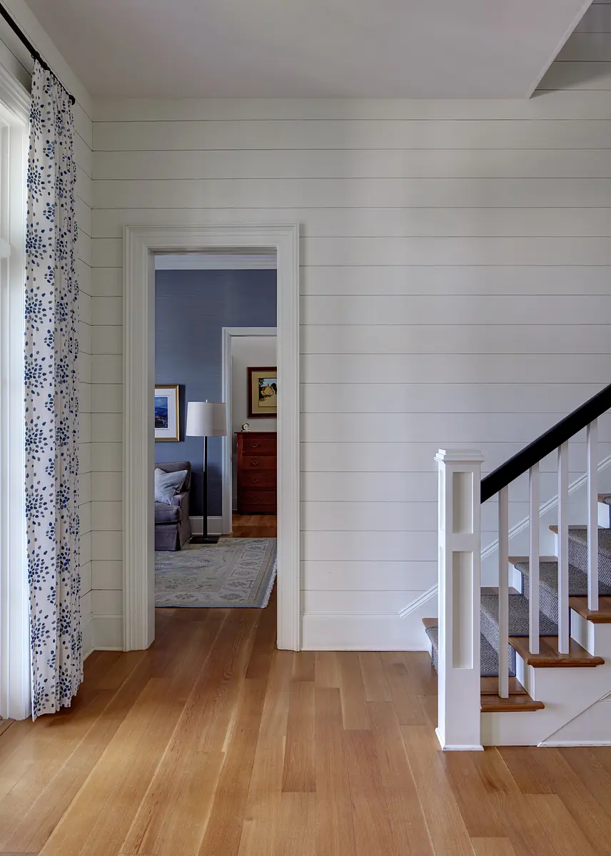 Hallway with hardwood flooring, white shiplap walls, staircase, and a patterned curtain.