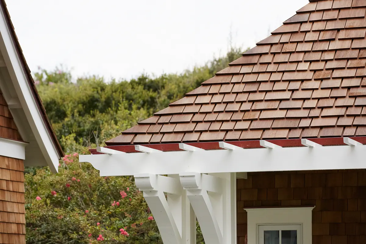 Exterior front view with gabled roof, wooden shingles, white trim, and greenery in the background.